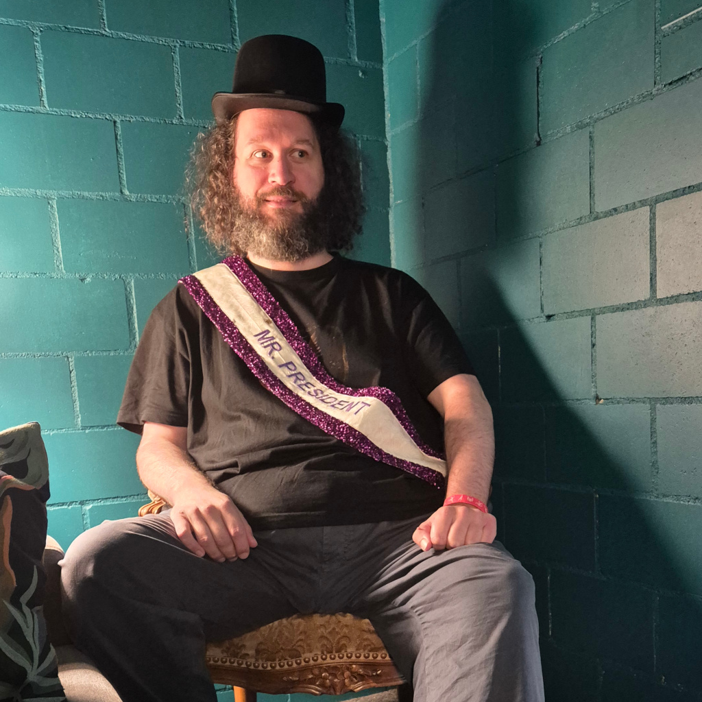 A photograph of a man with curly hair sitting in a chair, wearing a small top hat and a gaudy sash with the words Mr PResident on them.